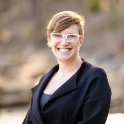 A woman with short hair in a black jacket and white glasses smiles broadly.